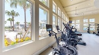 This expansive, single-floor fitness center offers a comprehensive workout environment designed to accommodate a wide range of exercise needs. The facility is architecturally distinguished by soaring vaulted and coffered ceilings, large multi-pane windows lining the perimeter walls, and an abundance of natural light that floods the open floor plan throughout the day. Ceiling fans are strategically placed overhead to ensure comfortable airflow across the space.

The main exercise area features a smooth, light-toned resilient floor with a curved design element that visually separates training zones. A full lineup of cardio equipment — including treadmills, ellipticals, stationary bikes, and rowing machines — is positioned along the windowed walls, offering views of palm trees and an outdoor pool area. An extensive strength training section houses a variety of Hoist cable machines and selectorized equipment with tan upholstery.

A dedicated functional training zone features turf flooring, TRX suspension systems, battle ropes, a mini trampoline, kettlebells, a dumbbell rack, and a wall-mounted training rig. A separate room includes spin bikes and a punching bag, accented by a teal feature wall with a decorative illustration. Wall-mounted televisions are positioned throughout, and built-in storage cabinetry provides organizational convenience.