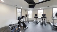 Located at 8115 Worthington Galena Rd in Westerville, Ohio, this single-floor fitness center is designed to provide a clean, functional workout environment. The space features a modern, contemporary aesthetic with crisp white walls, a stylish tray ceiling accented in dark trim, and recessed lighting complemented by a wall-mounted flat-screen TV — ideal for staying entertained during workouts. The flooring is finished with durable, textured gray material well-suited to high-traffic fitness use. Natural light flows generously through multiple large windows positioned around the room, offering views of the surrounding outdoor area and creating an open, energizing atmosphere. The facility is equipped with a well-rounded selection of cardio machines, including stationary bikes, ellipticals, and a multi-function pull-up and dip station, catering to a range of fitness routines. The layout is thoughtfully arranged to allow comfortable spacing between equipment, promoting ease of movement throughout the room. Ventilation is supported by ceiling vents, ensuring the space remains comfortable during use. Overall, this fitness center delivers a bright, well-equipped, and welcoming environment for users of all fitness levels.