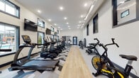 Located at 420 N Dean Rd in Auburn, Alabama, this single-floor fitness center is designed to deliver a comprehensive workout experience in a modern, well-appointed environment. The space is elongated and open, allowing for a generous layout that accommodates a wide range of equipment without feeling crowded. A row of TRUE-brand treadmills lines one side of the room, complemented by elliptical trainers, upright stationary bikes, and an air resistance fan bike. Strength training is supported by a cable machine system and a dumbbell rack positioned toward the rear of the space. The flooring combines light hardwood planks down the center lane with bold geometric-patterned carpet tiles in a black-and-white chevron and hexagon design, adding visual energy to the room. Multiple flat-screen TVs are mounted throughout, ensuring entertainment is accessible from any piece of equipment. Natural light filters in through rows of dark-framed windows positioned along both walls, with several offering direct views of an outdoor pool area. A bold orange accent wall near the entrance corridor makes a striking first impression and adds vibrancy to the otherwise neutral palette. Recessed and track lighting provide bright, even illumination throughout. A hand sanitizer station near the entry reflects a commitment to cleanliness and user convenience.