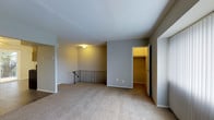 This single-floor residential space offers a clean, functional layout with a neutral color palette throughout. The main living area is finished with wall-to-wall beige carpeting, providing a warm and comfortable feel underfoot. Smooth white ceilings and light gray walls create a bright, open atmosphere, complemented by flush-mount ceiling lighting that evenly illuminates the space.

A notable architectural feature is the staircase landing, fitted with a decorative wrought iron railing that adds a subtle stylistic touch to the otherwise understated interior. The open railing design contributes to a sense of airiness within the space.

Adjacent to the main living area is a generously sized walk-in closet, visible through an open doorway and illuminated by interior lighting. The closet features a dedicated shelving and hanging rod system, offering ample built-in storage and organization options.

Additional doorways lead to further rooms or utility areas, suggesting a practical and well-connected floor plan. The overall design of the space prioritizes functionality and comfort, with consistent finishes and a straightforward layout that adapts well to a variety of uses. The neutral tones and carpeted flooring give the space a cozy, livable quality throughout.