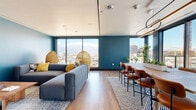 This thoughtfully designed single-floor space makes an immediate impression with its bold teal accent walls, warm wood-look flooring, and expansive floor-to-ceiling windows that frame breathtaking panoramic views of the surrounding mountains and city skyline. Natural light floods the interior throughout the day, enhancing the vibrant, contemporary atmosphere.

The open-plan layout seamlessly connects a generous lounge area with a dedicated dining space. The lounge is anchored by a large sectional sofa in deep charcoal, complemented by distinctive woven rattan egg chairs and a solid wood coffee table, all resting atop a textured area rug. A wall-mounted flat-screen television is positioned opposite the seating area, making the space equally suited for relaxation and entertainment.

Adjacent to the lounge, a substantial wooden dining table surrounded by leather-upholstered chairs serves as a central gathering point, illuminated by an elegant curved pendant light fixture. Lush potted plants add organic warmth throughout.

A decorative accent wall featuring a geometric line-art mural adds artistic interest near the entryway corridor, which leads to the main entry door. The space balances bold design choices with functional, welcoming furnishings, making it ideal for both social gatherings and everyday use.