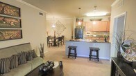 This single-floor residence showcases an open-concept layout that effortlessly connects the living room, dining area, and kitchen into one cohesive, flowing space. The living room features plush carpeting in neutral tones and is furnished with comfortable seating, anchored by a dark wood coffee table. Decorative accents, including sculptural floor vases, add character to the space.

The kitchen is a functional centerpiece, featuring warm honey-toned cabinetry, dark granite countertops, and a peninsula with bar-height seating for two. Pendant lighting hangs above the peninsula, providing both task lighting and a stylish focal point. A full suite of appliances, including a black refrigerator, completes the kitchen. Adjacent to the kitchen, the dining area accommodates a full dining table and chairs beneath an elegant chandelier, making it ideal for everyday meals or entertaining. A decorative wall medallion adds an artistic touch to this zone. Crown molding throughout the space lends a refined, polished finish, while the warm lighting palette creates an overall ambiance that feels cozy and welcoming.