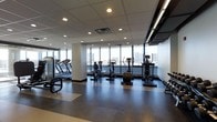 This well-appointed fitness center offers a thoughtfully designed workout environment suitable for a range of exercise routines. The space is anchored by an expansive bank of floor-to-ceiling windows that flood the interior with natural light while framing impressive panoramic city skyline views — a motivating backdrop for any workout.

The cardio equipment area is generously outfitted with multiple treadmills, upright and recumbent stationary bikes, and elliptical machines, all arranged to take full advantage of the window views. A dedicated free weights and stretching zone provides additional versatility for strength and flexibility training.

Underfoot, durable sport flooring in neutral tones provides both comfort and functionality. The dropped ceiling with recessed lighting ensures consistent, even illumination throughout the space, while HVAC vents are strategically positioned to maintain a comfortable climate. A wall-mounted display screen adds a modern, connected touch to the environment.

The overall ambiance is clean, contemporary, and professional, with a neutral color palette of whites, grays, and dark accents contributing to a focused and energizing atmosphere. The single-floor layout ensures easy accessibility, making this fitness center a practical and inviting space for daily use.