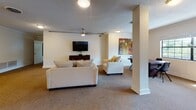 This single-story clubhouse in Jacksonville, Florida offers a well-appointed, open-concept layout designed for community gatherings and social events. The space is divided into two primary functional zones: a fully equipped kitchen area and a comfortable lounge, connected seamlessly by an open floor plan supported by decorative structural columns.

The kitchen is outfitted with light wood cabinetry extending to the ceiling, stainless steel appliances including a range, microwave, and dishwasher, and a black refrigerator. Hardwood flooring anchors this area, which also features an extended bar counter with bar stool seating — ideal for casual dining or socializing. A sink and ample counter space make the kitchen fully functional for event catering needs.

The lounge area transitions to plush carpeted flooring and features a wall-mounted flat-screen television, a ceiling fan, and comfortable cream-toned sofas and accent chairs arranged around a glass-top coffee table. Neutral-toned walls, textured ceilings, and windows allowing natural light contribute to the welcoming ambiance. The overall design is practical yet inviting, making this clubhouse a versatile gathering space for residents.