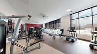 Located at 100 Pine St W in Wilson, NC, this fitness center offers a generously sized, single-floor exercise space designed to support a full range of workout routines. The layout is thoughtfully organized into distinct zones, seamlessly flowing between cardio and strength training areas across a curved floor plan with durable rubberized flooring throughout.

The cardio zone is lined with a row of treadmills and other aerobic machines positioned along floor-to-ceiling windows fitted with roller shades, allowing ample natural light to fill the space while offering outdoor views. Large ceiling fans overhead ensure comfortable air circulation throughout workouts.

The strength training area is equally well-appointed, featuring a comprehensive selection of selectorized cable machines, free-weight benches, and a full rack of dumbbells displayed along a mirrored wall that amplifies the sense of space. A dedicated functional training section includes a multi-use rack with TRX suspension trainers, medicine balls, and a stability ball, complemented by an Echelon rowing machine for added variety.

Bold red accent walls punctuate the otherwise clean white interior, adding energy and visual contrast. Wall-mounted televisions are positioned throughout for entertainment during workouts. The space strikes a balance between modern design and practical functionality, making it a comprehensive facility for fitness enthusiasts.