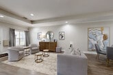 This well-appointed single-floor space offers an open-concept layout that seamlessly integrates multiple functional areas, creating a versatile and welcoming environment. The main living area is anchored by a stylish gray sofa, complemented by a pair of coordinating accent chairs and hexagonal gold-framed nesting tables atop a textured area rug. The neutral, warm-gray walls provide a refined backdrop for carefully curated artwork, including oversized abstract canvas pieces and decorative wall accents.

Recessed lighting throughout the space ensures consistent, ambient illumination, while a tray ceiling detail adds architectural interest and a sense of depth. Rich hardwood-style flooring flows continuously across the space, contrasting elegantly with the light tile found near the entry. Multiple windows dressed with layered drapes allow natural light to filter in, offering glimpses of the outdoors.

A dedicated workspace area features a substantial wood desk with seating, suited for productivity or consultation. Adjacent to this, a bar-height counter with upholstered stools connects to a compact kitchenette area equipped with cabinetry and appliances. The entry foyer provides a clean, welcoming transition into the space. Overall, the design balances comfort and functionality with a cohesive, contemporary-classic aesthetic.