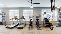 Located at 1004 Diamondback Dr in Sneads Ferry, NC, this fitness center spans a single floor and is organized into two dedicated exercise rooms, together offering a spacious and fully appointed workout facility. The main exercise room features patterned carpet tile flooring and is bathed in natural light from large windows fitted with blinds. Multiple ceiling fans keep the space comfortable, while pendant lighting adds a modern touch. This room houses an extensive array of cardio equipment, including treadmills, ellipticals, a stair climber, and a rowing machine, alongside a bold red power rack with free weights, a cable crossover machine, and a punching bag—catering to a wide range of fitness goals. A water station and wall-mounted TV are also present for added convenience. The second exercise room offers a change of surface with wood-look plank flooring and serves as a more flexible training space. It features spin bikes, stability balls, kettlebells, and a wall-mounted TV, with a mirrored wall and glass barn doors connecting it to the main room. Crown molding and neutral gray walls throughout both rooms lend a clean, polished aesthetic, while abundant natural light and thoughtful equipment arrangement create an inviting and functional environment for all fitness levels.