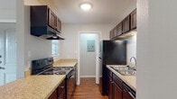 This single-floor unit offers a well-organized layout that balances functionality and comfort across its living spaces. The galley-style kitchen is a standout feature, equipped with dark espresso cabinetry that provides ample upper and lower storage, granite-style countertops, black appliances including an electric range with overhead ventilation hood, a black refrigerator, and a stainless steel double-basin sink. Wood-look flooring runs throughout the kitchen, adding warmth to the space. A utility panel is accessible through a door at the end of the kitchen corridor.

The open living area extends naturally from the kitchen, anchored by rich hardwood-style flooring and neutral gray walls that create a clean, versatile backdrop. A ceiling fan overhead adds comfort, while a sliding glass door at one end of the room allows natural light to filter in and provides outdoor access.

The bedroom is a comfortable, carpeted room featuring a ceiling fan with integrated light fixture and a window with blinds that introduces additional natural light. The room offers a quiet, restful atmosphere with ample wall space.

The bathroom is finished with consistent design elements, including dark espresso cabinetry beneath a granite-style countertop, a single-basin sink, a large wall mirror, and wood-look flooring, tying together the cohesive aesthetic found throughout the unit.