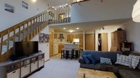 This two-story space is defined by its dramatic double-height ceilings on the main level, creating an open, airy atmosphere that immediately draws the eye upward. A wooden staircase with baluster railing leads to an upper loft level, which overlooks the main living area below through an open railing — a standout architectural feature that adds depth and visual interest to the interior.

The main level seamlessly integrates the living and kitchen areas into a cohesive open-concept layout. The living space is carpeted and comfortably accommodates a sofa and media console, offering a relaxed, functional setup. Recessed and track lighting throughout ensures the space remains well-lit.

The kitchen features warm honey-toned cabinetry, a central island with bar-height seating for casual dining, and standard appliances including a refrigerator and stovetop. A door adjacent to the kitchen provides access to additional rooms or storage beyond the main living space. A closet area is also visible near the living space, offering practical storage solutions. The overall ambiance is casual and functional, with warm tones and an efficient use of vertical space throughout.