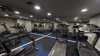 Located in Anderson, this well-equipped fitness center offers a comprehensive workout environment on a single floor. The space is designed with functionality and aesthetics in mind, featuring a striking blue and gray geometric-patterned carpet that adds a modern, energetic feel to the room.

The cardio section is prominently outfitted with multiple Landice treadmills, providing users with high-quality running options, alongside an elliptical machine for low-impact training. A multi-station strength training machine anchors the weightlifting area, offering a wide variety of resistance exercises suitable for full-body workouts. Flat workout benches are also available for additional strength training versatility.

Floor-to-ceiling mirrors line a full wall of the space, visually expanding the room and allowing users to monitor their form during exercise. Recessed lighting throughout the ceiling provides bright, even illumination across the entire floor. The walls are finished in a neutral gray tone, contributing to a clean and focused atmosphere.

A hand sanitizer station and fire safety equipment are mounted near the entry area, reflecting attention to hygiene and safety. A glass-paneled door with a dark frame connects the fitness center to an adjacent corridor with tile flooring, ensuring easy access. The overall design is practical, modern, and well-maintained.