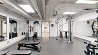 Located at 125 Wellington St N in Hamilton, Ontario, this exercise room is a comprehensive fitness facility spread across a single floor. The space is organized into distinct functional zones, each designed to support a well-rounded workout experience. The cardio area features light wood-look flooring, wall-to-wall mirrors that amplify the sense of space, a treadmill, an elliptical, and a mounted TV — all illuminated by bright, recessed panel lighting set against a clean white ceiling with exposed ductwork. Adjacent to this is a dedicated strength training zone with durable rubber flooring, a dual cable machine, a lat pulldown station, adjustable benches, and a full dumbbell rack, along with wall-mounted fans for ventilation. A separate open-concept room with the same light flooring provides flexible space suitable for stretching, yoga, or group exercise, and features a split-unit air conditioner for climate control. The facility also includes a kitchenette area equipped with a stone-topped counter, a sink, a refrigerator, and a standalone water cooler — offering convenient hydration and refreshment options. Throughout, the crisp white walls, consistent overhead lighting, and practical layout create a clean, functional, and motivating fitness environment.