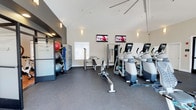 This fitness center offers a comprehensive workout environment designed to accommodate a range of exercise needs. The space is defined by its striking vaulted ceiling, which lends an open, airy quality to the room and enhances the sense of spaciousness throughout. Neutral white walls and subtle wall-mounted sconce lighting contribute to a clean, focused atmosphere conducive to exercise.

The cardio section features a row of elliptical machines and a rowing machine, positioned to face two wall-mounted flat-screen televisions, allowing users to stay entertained during their workouts. A cable strength machine and a weight bench are also present, providing options for resistance and strength training.

Near the entrance, a dedicated storage area organizes fitness accessories including stability balls in various sizes, resistance bands, and other small equipment, all neatly arranged on a rack unit. The frosted glass entry door, etched with "FITNESS," adds a polished, professional touch. Durable dark rubber flooring covers the entire space, offering safety and resilience underfoot. Natural light filters in through a window, further brightening this functional and well-appointed fitness facility.
