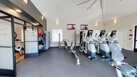 This fitness center offers a comprehensive workout environment designed to accommodate a range of exercise needs. The space is defined by its striking vaulted ceiling, which lends an open, airy quality to the room and enhances the sense of spaciousness throughout. Neutral white walls and subtle wall-mounted sconce lighting contribute to a clean, focused atmosphere conducive to exercise.

The cardio section features a row of elliptical machines and a rowing machine, positioned to face two wall-mounted flat-screen televisions, allowing users to stay entertained during their workouts. A cable strength machine and a weight bench are also present, providing options for resistance and strength training.

Near the entrance, a dedicated storage area organizes fitness accessories including stability balls in various sizes, resistance bands, and other small equipment, all neatly arranged on a rack unit. The frosted glass entry door, etched with "FITNESS," adds a polished, professional touch. Durable dark rubber flooring covers the entire space, offering safety and resilience underfoot. Natural light filters in through a window, further brightening this functional and well-appointed fitness facility.