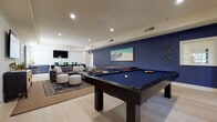 Located at 8445 Gardens Circle in Sarasota, Florida, this thoughtfully designed community clubhouse offers a diverse range of amenity spaces suited for both recreation and productivity. The grand entrance staircase sets an impressive tone, soaring to double-height ceilings beneath a sculptural chandelier, with bold teal wall art accenting the crisp white millwork and light hardwood floors. The expansive game and social lounge is a standout feature, anchored by a full-size pool table on navy felt, a shuffleboard table, and a large sectional seating area arranged beneath recessed lighting. A bold navy accent wall displaying coastal-themed artwork reinforces the relaxed Florida aesthetic. Adjacent to the lounge, a casual bar and dining area features subway tile, a beverage refrigerator, wall-mounted television, and bar-height seating with green metal chairs. The conference room accommodates a full complement of guests around a natural wood table, complemented by teal velvet chairs, blue-toned carpet, and a contemporary globe chandelier. A private business workstation nook with a marble-topped desk and garden-facing window provides a quiet workspace. A welcoming seating alcove with curated artwork serves as an ideal reception area. The covered outdoor veranda, lined with white columns and a beadboard ceiling, offers comfortable lounge seating with views of the surrounding landscape.