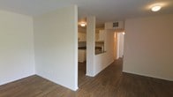 This single-floor residence offers a practical and open layout designed for comfortable everyday living. The main living area is finished with rich, dark hardwood flooring that extends throughout, providing a warm and cohesive aesthetic across the space. A partial dividing wall separates the living area from the kitchen, creating a sense of defined zones while maintaining an open, connected feel. The kitchen features white cabinetry, dark countertops, and a built-in range, all framed by a pass-through counter that opens toward the living space. Overhead lighting illuminates both areas, contributing to a bright and functional interior. A ceiling-mounted HVAC vent is visible, indicating a central air system. From the main living area, a hallway extends toward additional rooms, offering privacy and separation from the communal spaces. The walls throughout are finished in a clean, neutral white tone, providing a versatile backdrop that complements a variety of furnishing styles. Overall, the space presents a straightforward and well-organized floor plan suited for efficient single-level living.