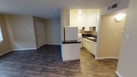 This single-floor residence showcases an open-concept design that seamlessly connects the kitchen and living areas, creating a cohesive and functional layout. The kitchen is well-appointed with white cabinetry extending to the ceiling, offering generous storage space. A mosaic tile backsplash in warm earth tones adds a decorative accent between the upper and lower cabinets, complementing the dark countertops. A peninsula with a dark countertop and white cabinetry below serves as a natural divider between the kitchen and the adjacent living space, providing additional prep or dining space. White appliances, including a refrigerator and range, are neatly integrated into the kitchen layout, alongside a sink with surrounding counter space. The living area features rich, dark hardwood-style flooring, while the kitchen transitions to a lighter wood-tone floor. A textured ceiling runs throughout the unit, and neutral-toned walls provide a versatile backdrop. The entry door is visible from the main living space, reflecting the unit's efficient, open floor plan.