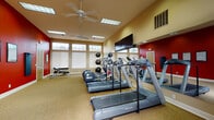 Located at 1133 North Grape Drive in Moses Lake, Washington, this dedicated fitness center offers a well-appointed, single-floor workout facility designed to support a full range of exercise routines. The space features bold red accent walls contrasted against warm beige tones, creating an energizing atmosphere that motivates activity. Ceiling fans and recessed fluorescent lighting ensure the room remains comfortable and well-illuminated throughout the day, while a row of transom windows along one wall allows natural light to filter in.

The cardio area is equipped with multiple treadmills and at least one elliptical machine, all positioned for ease of use and visibility to a wall-mounted flat-screen television. A full-length mirror wall visually expands the space and allows users to monitor their form during workouts. Strength training is supported by a set of medicine balls on a dedicated rack, a free weight station with a rack and adjustable bench, and additional functional training equipment. Informational charts and a corkboard are mounted on the walls for member reference.

Carpeted flooring covers the entire workout area, and the layout provides ample open floor space for stretching or bodyweight exercises. The room is accessed through a standard door and also includes an emergency exit, underscoring safety considerations. Overall, this fitness center presents a clean, functional, and motivating environment for users of all fitness levels.