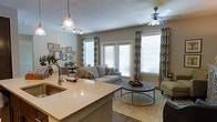 Located at 650 Enterprise Blvd in Charleston, South Carolina, this single-story residence offers a well-designed open-concept floor plan that seamlessly connects the kitchen, dining, and living areas. The kitchen features a generous island with light stone countertops, an undermount sink, and pendant lighting, making it both a functional workspace and a natural gathering point. Track lighting complements the pendant fixtures, ensuring the space is well-lit throughout the day. The living area is bright and inviting, anchored by large windows dressed with patterned drapes that allow ample natural light to fill the room. A ceiling fan overhead adds to the comfort of the space. The living room is furnished with a neutral-toned sofa, a layered area rug, and a round tiered coffee table, creating a relaxed yet polished atmosphere. Adjacent to the living area, a dedicated dining space is visible, offering a cohesive flow throughout the home. Dark hardwood flooring runs throughout, providing a rich contrast to the warm, neutral wall tones. The overall ambiance is modern and comfortable, with thoughtful design details that make this Charleston residence both functional and aesthetically refined.