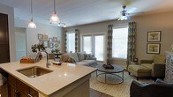 Located at 650 Enterprise Blvd in Charleston, South Carolina, this single-story residence offers a well-designed open-concept floor plan that seamlessly connects the kitchen, dining, and living areas. The kitchen features a generous island with light stone countertops, an undermount sink, and pendant lighting, making it both a functional workspace and a natural gathering point. Track lighting complements the pendant fixtures, ensuring the space is well-lit throughout the day. The living area is bright and inviting, anchored by large windows dressed with patterned drapes that allow ample natural light to fill the room. A ceiling fan overhead adds to the comfort of the space. The living room is furnished with a neutral-toned sofa, a layered area rug, and a round tiered coffee table, creating a relaxed yet polished atmosphere. Adjacent to the living area, a dedicated dining space is visible, offering a cohesive flow throughout the home. Dark hardwood flooring runs throughout, providing a rich contrast to the warm, neutral wall tones. The overall ambiance is modern and comfortable, with thoughtful design details that make this Charleston residence both functional and aesthetically refined.
