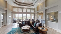 Located at 155 Lakeside Dr in Waxahachie, TX, this single-floor community clubhouse is designed to impress with its grand architectural details and welcoming atmosphere. The main lounge area commands attention with its dramatic coffered tray ceiling, soaring double-height walls, and an oversized ceiling fan that anchors the space. Floor-to-ceiling windows and large glass panels flood the interior with natural light while offering unobstructed views of the resort-style pool and outdoor patio area beyond.

The lounge is appointed with a generously sized brown leather sectional sofa, complemented by a vibrant teal and blue area rug and coordinating accent pillows. Wooden round coffee tables and side tables add a warm, natural touch. A separate casual dining area features sleek black bar-height tables and stools positioned directly in front of the pool-facing windows, ideal for socializing. Built-in shelving niches display tasteful décor, and a wall-mounted flat-screen TV provides an entertainment focal point.

Light hardwood-style flooring runs throughout, lending a clean and cohesive look. A beverage station with a credenza offers added convenience, while interior passageways with French doors connect to additional rooms, including what appears to be a private office or leasing area. The overall ambiance is polished yet relaxed, balancing upscale design with everyday functionality.