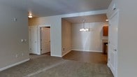 This single-floor space showcases an open-concept layout that seamlessly connects the living and dining areas, creating a cohesive and functional environment. The living area is finished with plush carpeting in a neutral tone, providing a comfortable and inviting atmosphere, while the dining area transitions to a smooth hard-surface floor, offering a practical distinction between the two spaces.

A pendant chandelier with multiple light fixtures hangs above the dining area, serving as a central decorative element and providing focused illumination. Recessed lighting supplements the overall brightness of the space. The textured ceiling adds subtle dimension throughout.

Warm beige walls run consistently across both areas, contributing to the space's unified and calm aesthetic. White trim and door casings provide clean contrast against the neutral walls. Two white panel doors are visible, leading to additional areas of the unit. A wall vent ensures climate control comfort.

To one side, warm wood-toned cabinetry is partially visible, hinting at an adjacent kitchen space. The overall layout is straightforward and well-proportioned, making it an adaptable space suited to a variety of furnishing arrangements.