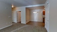 This single-floor space showcases an open-concept layout that seamlessly connects the living and dining areas, creating a cohesive and functional environment. The living area is finished with plush carpeting in a neutral tone, providing a comfortable and inviting atmosphere, while the dining area transitions to a smooth hard-surface floor, offering a practical distinction between the two spaces.

A pendant chandelier with multiple light fixtures hangs above the dining area, serving as a central decorative element and providing focused illumination. Recessed lighting supplements the overall brightness of the space. The textured ceiling adds subtle dimension throughout.

Warm beige walls run consistently across both areas, contributing to the space's unified and calm aesthetic. White trim and door casings provide clean contrast against the neutral walls. Two white panel doors are visible, leading to additional areas of the unit. A wall vent ensures climate control comfort.

To one side, warm wood-toned cabinetry is partially visible, hinting at an adjacent kitchen space. The overall layout is straightforward and well-proportioned, making it an adaptable space suited to a variety of furnishing arrangements.