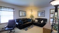 This three-floor property offers a comfortable and functional layout across its multiple levels. The living room, as seen, is a welcoming space anchored by a large dark leather sectional sofa and a matching loveseat, arranged to encourage conversation and relaxation. A patterned area rug in muted earth tones grounds the seating arrangement, while carpeted flooring adds warmth underfoot. The room is illuminated by a chandelier-style pendant light, complemented by a floor lamp that casts a soft, ambient glow. Decorative wall mirrors and framed artwork add character to the light-colored walls, and a potted plant in the corner introduces a touch of greenery. Natural light filters in through the windows, enhancing the overall brightness of the space. The neutral wall tones and understated décor create a calm, livable atmosphere. With three floors of living space, this property provides ample room for various uses, offering flexibility and generous square footage spread vertically across its levels.
