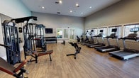 This fitness center offers a comprehensive workout environment designed to accommodate a variety of exercise routines. The space features hardwood-style flooring throughout, contributing to a clean and polished aesthetic. Neutral gray walls and recessed lighting create a bright, energizing atmosphere conducive to physical activity.

The cardio section is well-appointed with multiple treadmills and elliptical machines, each equipped with individual monitors for entertainment or workout tracking. These are positioned along the windowed wall, which features slatted blinds allowing natural light to filter into the space.

The strength training area includes a multi-functional cable machine, an adjustable workout bench, and a dedicated free weight station with a full rack of dumbbells. Exercise balls in varying sizes are also available, visible near the mirrored wall, which helps visually expand the space and allows users to monitor their form during workouts.

A wall-mounted flat-screen television is positioned near the free weight area, adding a convenient entertainment option. The combination of cardio equipment, strength machines, and free weights makes this fitness center a versatile facility suitable for a wide range of fitness levels and training goals.