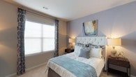 The bedroom presents a calm and inviting atmosphere, anchored by soft lavender-gray walls that complement the neutral carpeted flooring. A large window dressed with patterned curtains and white horizontal blinds allows generous natural light to filter into the room, creating a bright and airy feel. The tufted white upholstered headboard serves as a focal point, lending a touch of elegance to the space. Coordinating bedding in soft aqua and cream tones adds layers of texture and visual interest. A dark wood nightstand with a classic table lamp sits alongside the bed, providing functional bedside lighting. Abstract wall art in muted tones adds a decorative accent above the headboard. The smooth white ceiling features a centrally placed air vent, reflecting a clean and functional design. Overall, the bedroom balances comfort and style through its cohesive color palette, soft furnishings, and well-considered layout, offering a restful and welcoming environment suited for relaxation.