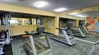 This fitness center offers a focused and fully equipped environment designed to support a well-rounded workout routine. The space is outfitted with multiple Matrix-brand treadmills positioned for comfortable use, providing ample room between machines for safe and unrestricted exercise. Along the walls, large floor-to-ceiling mirrors extend the visual depth of the room while allowing users to monitor their form during workouts. A dedicated strength training and cable machine station is visible along the mirrored back wall, offering options for resistance and functional training in addition to cardiovascular exercise. The flooring features a durable, patterned carpet suitable for a high-traffic fitness environment, with colorful exercise mats available for stretching or floor-based workouts. Recessed flush-mount ceiling lights provide consistent, even illumination throughout the space, ensuring a bright and energizing atmosphere. The warm-toned walls contribute to an inviting ambiance, while the layout is thoughtfully organized to maximize usability. Informational and motivational signage is displayed on the walls, adding a practical and encouraging touch to the overall environment. This single-floor fitness facility is a convenient and comprehensive amenity for those seeking a dedicated space for their health and wellness needs.
