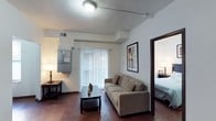 This single-floor space showcases a well-appointed living area anchored by rich, dark hardwood flooring that extends throughout the main areas, creating a warm and cohesive aesthetic. The living room is furnished with a neutral-toned sofa accented by patterned throw pillows, complemented by a dark wood coffee table that grounds the space. A sliding glass door dressed with vertical blinds serves as the focal point of the living area, allowing natural light to filter in while providing access to the exterior. A framed piece of wall art adds a decorative touch above the seating area. The textured ceiling and neutral white walls contribute to a clean, understated backdrop throughout. A utility panel is mounted on the wall near the entry, offering practical accessibility. Through an open doorway, the bedroom is visible, featuring matching hardwood floors, a neatly made bed, and a bedside lamp that echoes the warm lighting found in the living space. The overall ambiance is modest yet functional, with consistent finishes and a straightforward layout designed for comfortable everyday living.