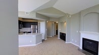 This single-floor space showcases an open-concept layout that seamlessly integrates the kitchen and living areas, creating a cohesive and functional environment. The vaulted ceilings are a standout architectural feature, adding a sense of vertical spaciousness and airiness throughout the main living area. Neutral gray walls complement the light-toned carpeting that extends across the living space, creating a clean, modern aesthetic.

The kitchen is appointed with dark espresso cabinetry that contrasts sharply against a light tile backsplash. Stainless steel appliances, including a refrigerator and range, add a contemporary touch, while the granite-topped peninsula island provides additional workspace and serves as a natural divider between the kitchen and living areas. The peninsula's curved edge adds a subtle design detail.

Beyond the main living space, a hallway leads to additional rooms, with glimpses of a built-in shelving unit and additional dark cabinetry visible in an adjacent nook or secondary space. HVAC vents are integrated into the upper walls, ensuring climate control throughout. The overall ambiance is modern, practical, and well-organized, with consistent finishes tying the space together cohesively.