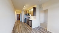 This single-floor unit showcases a clean, contemporary design with warm wood-tone cabinetry and durable luxury vinyl plank flooring that flows throughout the main living areas. The kitchen serves as a functional centerpiece, featuring stainless steel appliances including a refrigerator, electric range, built-in microwave, and dishwasher. Dark-toned lower cabinets are paired with lighter upper cabinetry, complemented by white quartz countertops and a classic subway tile backsplash. Track lighting overhead ensures the kitchen is well-lit and practical. Adjacent to the kitchen, the open living area benefits from natural light streaming through a large window, and is equipped with a wall-mounted mini-split heating and cooling unit for year-round comfort. A long hallway connects the main living spaces to the private areas of the unit. The bathroom is spacious and thoughtfully designed, featuring a wide vanity with a white countertop, dark cabinetry with ample storage, an overhead cabinet, and grab bars indicating accessibility-conscious design. Light-toned tile flooring completes the bathroom's fresh, neutral aesthetic. The overall ambiance of the unit is bright, modern, and move-in ready.