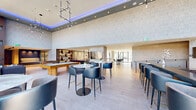 This sophisticated amenity lounge is designed to impress, combining thoughtful layout with high-end finishes throughout its single-floor footprint. The space is anchored by dramatic floor-to-ceiling windows that frame sweeping urban skyline views, flooding the interior with natural light and creating a seamless connection between indoors and the cityscape beyond. Soaring double-height ceilings, accented by subtle blue LED lighting along the upper perimeter, lend an upscale, contemporary atmosphere.

The lounge is divided into several distinct zones. A spacious social area features plush sectional sofas, a large flat-screen TV, and a sleek built-in entertainment wall with illuminated shelving flanking a linear fireplace — ideal for relaxed gatherings. An adjacent game room includes both a pool table and a foosball table, complemented by backlit display shelving and wall-mounted screens.

The dining and bar area offers high-top marble-topped tables with stylish black and gold accent chairs, along with bar seating along the windows to maximize views. A fully equipped catering kitchen with dark wood cabinetry, grey subway tile, and stainless steel appliances supports event hosting. Light wood-look tile flooring unifies the entire space, completing its refined, contemporary aesthetic.