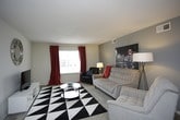 This single-floor residence offers a thoughtfully designed layout that balances comfort and functionality across its living spaces. The living room serves as the heart of the home, featuring a contemporary tufted sofa, mid-century modern accent chairs, and a bold black-and-white geometric area rug. Neutral gray walls and wood-look flooring lend a modern aesthetic, while a large window dressed with vibrant red curtains fills the room with natural light.

The galley-style kitchen is efficiently organized, offering ample cabinetry, dark countertops, a double-basin sink, and black appliances including a range and refrigerator. Wood-look flooring continues here, complementing the clean, practical design.

The residence includes multiple bedrooms, each with its own character. The primary bedroom features a gray accent wall, plush carpeting, a queen-sized bed, and a bright window that invites natural light. A second bedroom echoes a similar style with a gray accent wall and built-in closet storage. A third bedroom offers a lighter, neutral palette with a dedicated workspace, a window with blinds, and carpeted flooring — ideal as a guest room or home office.

The bathroom is clean and straightforward, with wood-look flooring, a vanity with dark countertop, and a tub-shower combination. This residence presents a cohesive, modern living environment with practical amenities throughout.