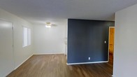 This single-floor residence offers a clean and functional layout designed with simplicity and contrast in mind. The main living area is anchored by warm, medium-toned hardwood-style flooring that runs throughout, providing a cohesive foundation for the space. A striking dark navy-blue accent wall serves as a bold focal point, creating a strong visual contrast against the crisp white walls that line the rest of the room. The textured popcorn ceiling adds a classic touch, while a central ceiling-mounted light fixture provides ambient illumination to the living area. The open layout allows for flexible furniture arrangement and a sense of spaciousness. A glimpse into the adjacent area reveals white cabinetry, suggesting a connected kitchen or utility space that maintains the home's clean, neutral aesthetic. Natural light filters into the space through the windows, complementing the overall bright and airy feel. The combination of the rich flooring, bold accent wall, and white trim and walls gives this residence a modern yet approachable character, well-suited for comfortable everyday living.