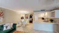 Located at 5151 Richmond Ave in Houston, TX, this single-floor unit offers a well-designed open-concept layout that seamlessly connects the living, dining, and kitchen areas. The living room is anchored by a plush green tufted velvet sofa, crown molding, and a large abstract canvas, with natural light filtering in through vertical blinds. Adjacent to the living space is a compact dining area featuring a glass-top table and contemporary white chairs beneath a pendant light. The kitchen is thoughtfully appointed with white cabinetry, a tile backsplash, a built-in wine rack display, light tile countertops, and a full suite of appliances including a range, microwave, dishwasher, and refrigerator. A peninsula counter separates the kitchen from the living and dining spaces, creating a natural flow throughout. The bedroom is a comfortable retreat with rich hardwood flooring, crown molding, a ceiling fan, a tufted upholstered bed frame, and a full wall of mirrored sliding closet doors that amplify the sense of space. Warm, neutral tones are carried throughout the unit, complemented by tile flooring in the main living areas. The overall ambiance is modern and inviting, blending practical functionality with thoughtful design details.