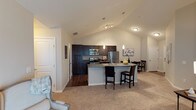 This single-floor residence showcases an open-concept layout that seamlessly integrates the living, dining, and kitchen areas into one cohesive space. A standout architectural feature is the vaulted ceiling, which peaks dramatically over the main living area, adding a sense of volume and airiness throughout.

The kitchen is outfitted with rich, dark espresso cabinetry that extends to the upper walls, providing generous storage capacity. Stainless steel appliances, including a refrigerator, range, and built-in microwave, lend a modern, polished aesthetic. The kitchen peninsula serves as both a functional workspace and a casual dining spot, equipped with a built-in sink and seating for two with dark upholstered bar stools. Pendant lighting above the peninsula adds focused illumination and a refined design touch, complemented by a track lighting fixture overhead.

The living area transitions smoothly from the kitchen, featuring plush neutral carpeting that contrasts with the hardwood-style flooring in the kitchen zone. The dining area, positioned adjacent to the kitchen, offers space for a small table and chairs. Neutral wall tones and white trim throughout contribute to a clean, contemporary ambiance.