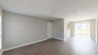 This single-story unit is designed with a practical, open layout that connects the main living areas seamlessly. The living room is spacious, featuring dark hardwood-style flooring, neutral gray walls, and clean white baseboards throughout. A tile-surround wood-burning fireplace serves as a focal point, accompanied by a window that draws in natural light. The open-plan layout connects the living area to the kitchen, which is equipped with gray cabinetry, a white electric range, an overhead range hood, and a peninsula with a light stone-style countertop. A sliding glass door adjacent to the kitchen provides direct access to an outdoor area and allows additional natural light into the space. The bedrooms are finished with plush carpet and ceiling fans with integrated lighting. The primary bedroom includes direct access to a bathroom featuring a wide vanity with a speckled stone-style countertop, a large mirror, and ample cabinet storage. The bathroom also includes a full-size soaking tub with a shower rod. A second bathroom mirrors this design. Textured ceilings are present throughout the unit.