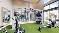 Located at 2525 Paseo Park Rd in Orlando, Florida, this single-story fitness center is designed to support a comprehensive range of workout modalities in a bright, contemporary setting. The facility is divided into two distinct areas that together create a well-rounded training environment.

The main training floor features artificial turf throughout, providing an ideal surface for functional fitness and high-intensity workouts. This zone is equipped with air bikes, rowing machines, a StairMaster bench, and a suspended rig system outfitted with TRX suspension trainers, battle ropes, and storage for medicine balls and kettlebells. Full-length mirrors line the walls, amplifying the sense of space and allowing users to monitor their form. A coffered ceiling with a statement sputnik chandelier and warm accent lighting adds a polished, upscale aesthetic to the space.

A glass-enclosed spin studio adjoins the main floor, housing multiple cycling bikes arranged before a large-format display screen. The light wood flooring and crisp white walls give this room a clean, focused atmosphere, while strategically placed speakers support an immersive cycling experience.

Floor-to-ceiling windows along the perimeter of the facility flood both zones with abundant natural light and offer open views of the surrounding outdoor landscape, creating an energizing and welcoming workout environment throughout the day.