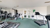 Located at 65 Villa Rd in Greenville, South Carolina, this fitness center offers a well-equipped exercise space designed to support a range of workout routines. The room features clean white walls, recessed lighting, and green rubber-topped flooring that provides both cushioning and durability underfoot. A full wall of mirrors along one side visually expands the space and aids in maintaining proper form during workouts. The cardio section is well-represented with multiple treadmills, a recumbent bicycle, an upright stationary bike, and an elliptical trainer. Strength training options include a cable pulley machine and several selectorized weight machines, offering a comprehensive range of resistance exercises. The space benefits from natural light streaming in through large glass double doors, which open to an outdoor deck area, creating a bright and airy atmosphere. A secondary set of glass double doors provides additional entry and connectivity to the surrounding property. Recessed ceiling lights and overhead spotlights ensure the room remains well-lit throughout the day. The overall layout is practical and efficient, making it a functional fitness facility for residents of the community.