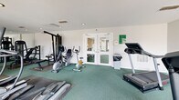 Located at 65 Villa Rd in Greenville, South Carolina, this fitness center offers a well-equipped exercise space designed to support a range of workout routines. The room features clean white walls, recessed lighting, and green rubber-topped flooring that provides both cushioning and durability underfoot. A full wall of mirrors along one side visually expands the space and aids in maintaining proper form during workouts. The cardio section is well-represented with multiple treadmills, a recumbent bicycle, an upright stationary bike, and an elliptical trainer. Strength training options include a cable pulley machine and several selectorized weight machines, offering a comprehensive range of resistance exercises. The space benefits from natural light streaming in through large glass double doors, which open to an outdoor deck area, creating a bright and airy atmosphere. A secondary set of glass double doors provides additional entry and connectivity to the surrounding property. Recessed ceiling lights and overhead spotlights ensure the room remains well-lit throughout the day. The overall layout is practical and efficient, making it a functional fitness facility for residents of the community.