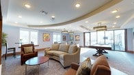 This single-floor community lounge is a well-appointed gathering space that blends relaxed comfort with entertainment amenities. The room's most striking architectural feature is its curved tray ceiling, which adds a dynamic, sculptural element to the interior and helps define distinct zones within the open layout. Recessed lighting throughout the ceiling is complemented by decorative pendant fixtures with warm metallic finishes, ensuring well-distributed illumination across the space.

The seating area is thoughtfully arranged, featuring a curved upholstered sofa, plush armchairs in neutral and warm brown tones, and patterned accent pillows that add visual interest. A round side table anchors one corner of the seating arrangement, contributing to an inviting, conversational atmosphere. The flooring transitions between hardwood and a decorative area rug with an organic swirl pattern beneath the primary seating cluster.

Toward the perimeter, a full-sized pool table serves as a central entertainment feature, positioned near floor-to-ceiling glass doors and large windows that flood the space with natural light and offer views of the outdoors. Colorful framed artwork adorns the walls, adding a casual, lively energy throughout.
