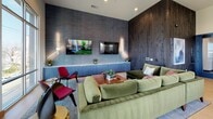 Located at 4600 Ross Ave in Dallas's vibrant Ross Avenue corridor, this single-floor space delivers a compelling blend of contemporary design and urban sophistication. The main lounge area commands attention with soaring accent walls clad in dark, richly textured wood paneling that reaches toward the ceiling, creating a dramatic focal point. Complementing this is a slate-blue textured feature wall housing a built-in media cabinet with subtle underlighting and dual wall-mounted flat-screen TVs, ideal for entertainment and gathering. Plush sage-green velvet sectional seating, vibrant crimson accent chairs, and a warm wood coffee table furnish the space with both comfort and visual interest. Light wide-plank hardwood flooring runs throughout, grounding the interior with warmth. A bar-height counter with wooden stools lines the perimeter near a kitchenette area, providing casual seating alongside a stainless steel sink and dishwasher. Floor-to-ceiling windows and a bank of casement-style glazing flood the space with natural light while framing panoramic views of the Dallas skyline and a neighboring historic landmark building. Curated wall art and thoughtful accent lighting complete the modern, community-oriented ambiance of this well-appointed space.