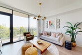 This thoughtfully designed single-floor residence blends contemporary architecture with a warm, livable aesthetic. Wide-plank hardwood flooring runs throughout, unifying the space from the welcoming entry hallway to the open-concept living and kitchen areas beyond.

The kitchen is a standout feature, anchored by a large waterfall-edge island with seating for multiple guests, illuminated by elegant glass pendant lights. Glossy white cabinetry, a herringbone tile backsplash, stainless steel appliances, and a professional-grade range make it as functional as it is visually striking.

The living area is bathed in natural light through expansive floor-to-ceiling windows that frame sweeping outdoor views and open to a balcony, creating a seamless indoor-outdoor connection. A modern chandelier with brass accents adds a refined touch overhead.

The bedroom continues the floor-to-ceiling window theme, offering serene treetop views and a bold blue accent wall that adds depth and character. An adjoining walk-in closet provides generous built-in shelving and hanging storage.

A dedicated home office features botanical wallpaper and ample shelving, offering a quiet, inspiring workspace. The bathroom is finished with large-format subway tile, a glass-enclosed walk-in shower, and warm neutral tones throughout, completing this polished and well-appointed residence.