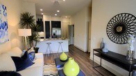 This contemporary single-floor residence showcases a thoughtfully designed open-concept layout that seamlessly connects the living and kitchen areas, creating a sense of spaciousness and flow. Rich, dark hardwood flooring runs throughout, anchoring the space with warmth and sophistication.

The kitchen is a standout feature, outfitted with dark cabinetry that contrasts elegantly against light stone countertops and a classic subway tile backsplash. A stainless steel refrigerator with an ice and water dispenser adds a modern touch, while a peninsula with bar seating — accommodating sleek metallic stools — provides a casual dining and entertaining option. Recessed lighting illuminates the kitchen and living areas efficiently.

The open living area flows naturally from the kitchen, offering ample room for comfortable furnishings. Neutral, warm-toned walls provide a versatile backdrop throughout the space. A side console table adds a functional yet decorative element near the entryway corridor.

The layout includes a hallway that leads to additional areas of the residence, suggesting a well-organized floor plan. Overall, the space presents a clean, modern aesthetic with a balanced mix of dark and light tones, making it a stylish and functional living environment.