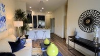 This contemporary single-floor residence showcases a thoughtfully designed open-concept layout that seamlessly connects the living and kitchen areas, creating a sense of spaciousness and flow. Rich, dark hardwood flooring runs throughout, anchoring the space with warmth and sophistication.

The kitchen is a standout feature, outfitted with dark cabinetry that contrasts elegantly against light stone countertops and a classic subway tile backsplash. A stainless steel refrigerator with an ice and water dispenser adds a modern touch, while a peninsula with bar seating — accommodating sleek metallic stools — provides a casual dining and entertaining option. Recessed lighting illuminates the kitchen and living areas efficiently.

The open living area flows naturally from the kitchen, offering ample room for comfortable furnishings. Neutral, warm-toned walls provide a versatile backdrop throughout the space. A side console table adds a functional yet decorative element near the entryway corridor.

The layout includes a hallway that leads to additional areas of the residence, suggesting a well-organized floor plan. Overall, the space presents a clean, modern aesthetic with a balanced mix of dark and light tones, making it a stylish and functional living environment.