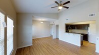 This single-floor residence offers an open and airy layout designed for comfortable everyday living. The main living area features warm hardwood-style flooring that extends throughout, creating a cohesive and inviting atmosphere. Vaulted ceilings amplify the sense of space, while a ceiling fan with integrated lighting provides both comfort and illumination. The living area flows seamlessly into an adjoining dining or flex space, lit by a classic pendant light fixture, adding a welcoming ambiance to the open floor plan. The kitchen is accessible via a convenient pass-through counter with a breakfast bar overhang, ideal for casual dining or entertaining. Dark espresso-toned cabinetry is paired with light granite countertops, and stainless steel appliances complete the functional and stylish kitchen setup. A doorway visible from the main living area leads to additional rooms, suggesting well-organized private spaces beyond. The clean white walls and neutral color palette throughout create a bright, move-in-ready environment. The thoughtful layout and architectural details — including the vaulted ceiling and open sightlines — make this a practical and visually appealing single-floor living space.