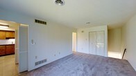 This single-floor space presents a clean, functional layout designed with simplicity and practicality in mind. The interior is finished in soft, neutral tones throughout, creating a calm and versatile atmosphere well-suited to a variety of uses.

The main room is generously sized, offering an open and airy feel enhanced by plush wall-to-wall carpeting in a muted gray tone. The room features white walls and a smooth ceiling with centrally located ventilation, ensuring comfortable climate control year-round. A dedicated HVAC return vent and additional floor-level vents further support efficient air circulation throughout the space.

Built-in bifold closet doors along one wall provide ample storage, blending seamlessly with the room's understated aesthetic. Multiple electrical outlets are positioned conveniently around the perimeter, adding to the room's practicality. A doorway leads to an adjacent area, suggesting additional connected rooms that complement the overall floor plan.

The overall ambiance is bright, neutral, and move-in ready, offering a blank canvas that lends itself well to personalization. The straightforward design, combined with thoughtful built-in features, makes this a comfortable and adaptable single-floor space.