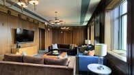 This distinguished lounge exudes timeless elegance through its masterful blend of classic architectural detailing and carefully curated furnishings. The walls are clad in rich, dark wood paneling that extends from floor to ceiling, lending the space a warm and stately character. Intricate crown molding and ornamental plasterwork frame the ceiling, which is accented with a deep teal inset that adds a refined touch of color and visual depth. Dual chandeliers with warm-toned shades cast a soft, welcoming glow throughout the room, complementing the ambient lighting provided by classic table lamps. The seating arrangement features a mix of plush velvet sofas in deep brown and cool blue tones, arranged to encourage both intimate conversation and relaxed lounging. A lacquered coffee table serves as a central anchor, while side tables provide practical elegance throughout. A large flat-screen television is mounted on one wall, adding a modern convenience without disrupting the room's traditional aesthetic. The overall ambiance is one of a private club or upscale hospitality setting — sophisticated, inviting, and richly detailed at every turn.